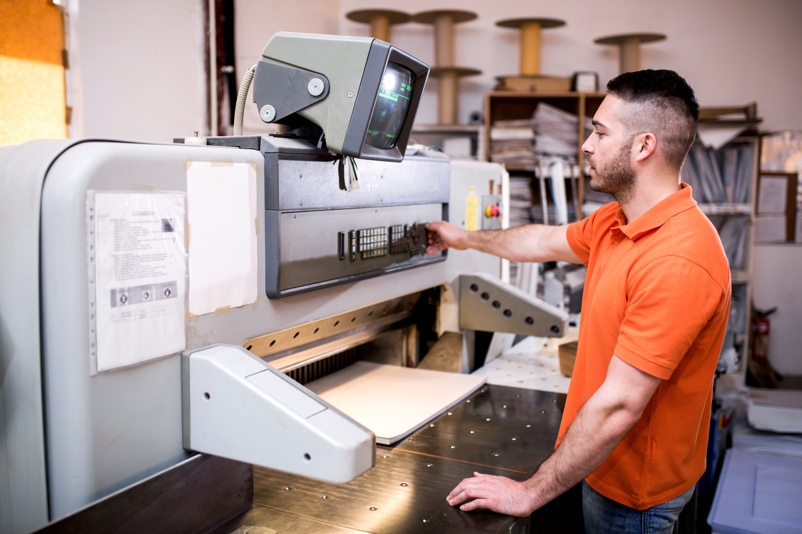 Man operating a large industrial printing machine in a print shop, adjusting settings on the control panel.