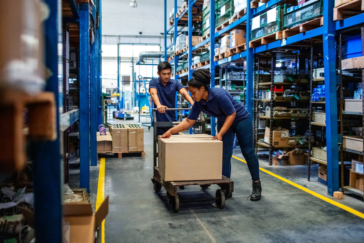 Two warehouse workers efficiently managing packaging and logistics, moving a box on a cart through organized shelves in a distribution center.